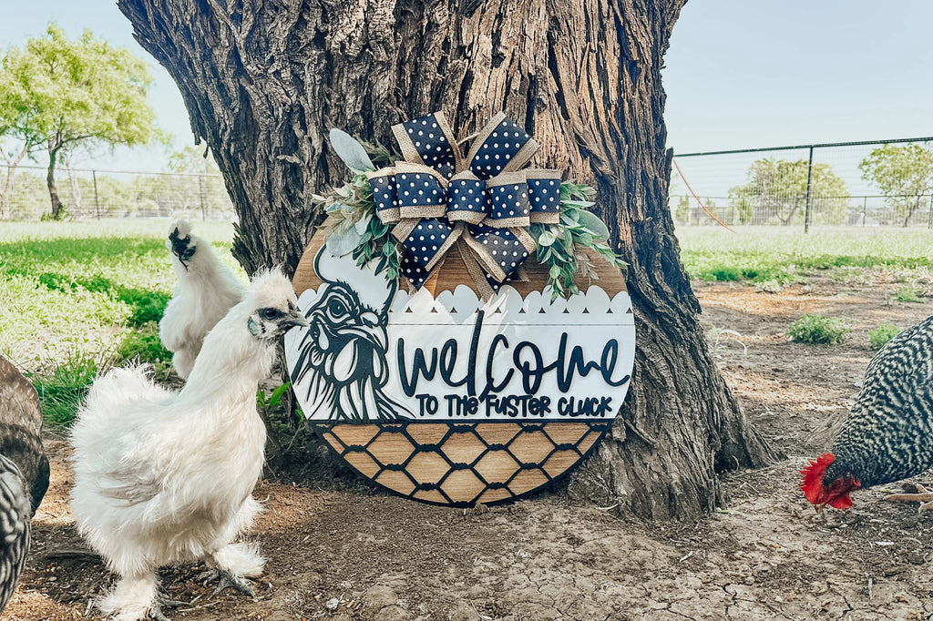 A round wooden sign with a chicken design, greenery, and a black-and-white polka dot bow reads Welcome to the Fuster Cluck. Chickens stand nearby under a tree in a fenced outdoor area.