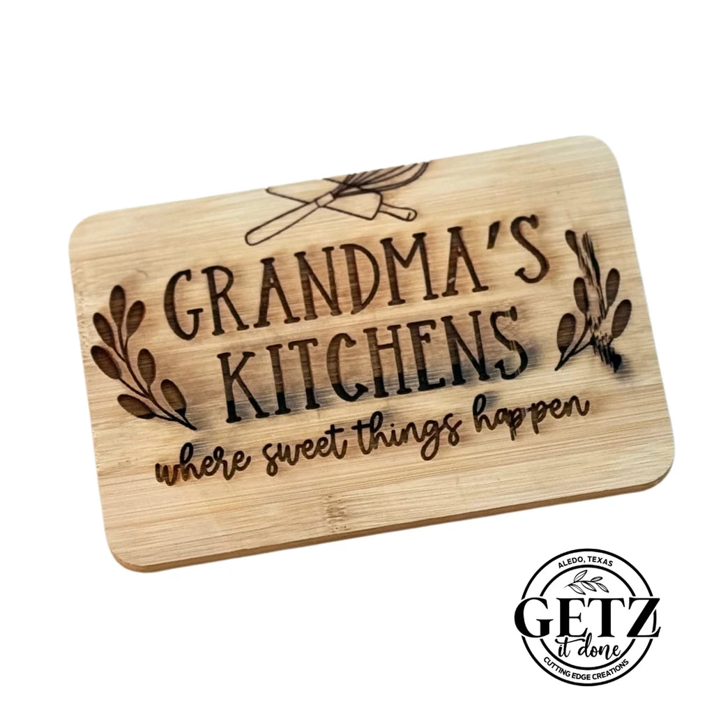 A wooden cutting board engraved with GRANDMAS KITCHEN where sweet things happen featuring kitchen utensils and laurel branch designs. The bottom right displays a black circular logo with text: GETZ done and Aledo, Texas with a cutting edge design.