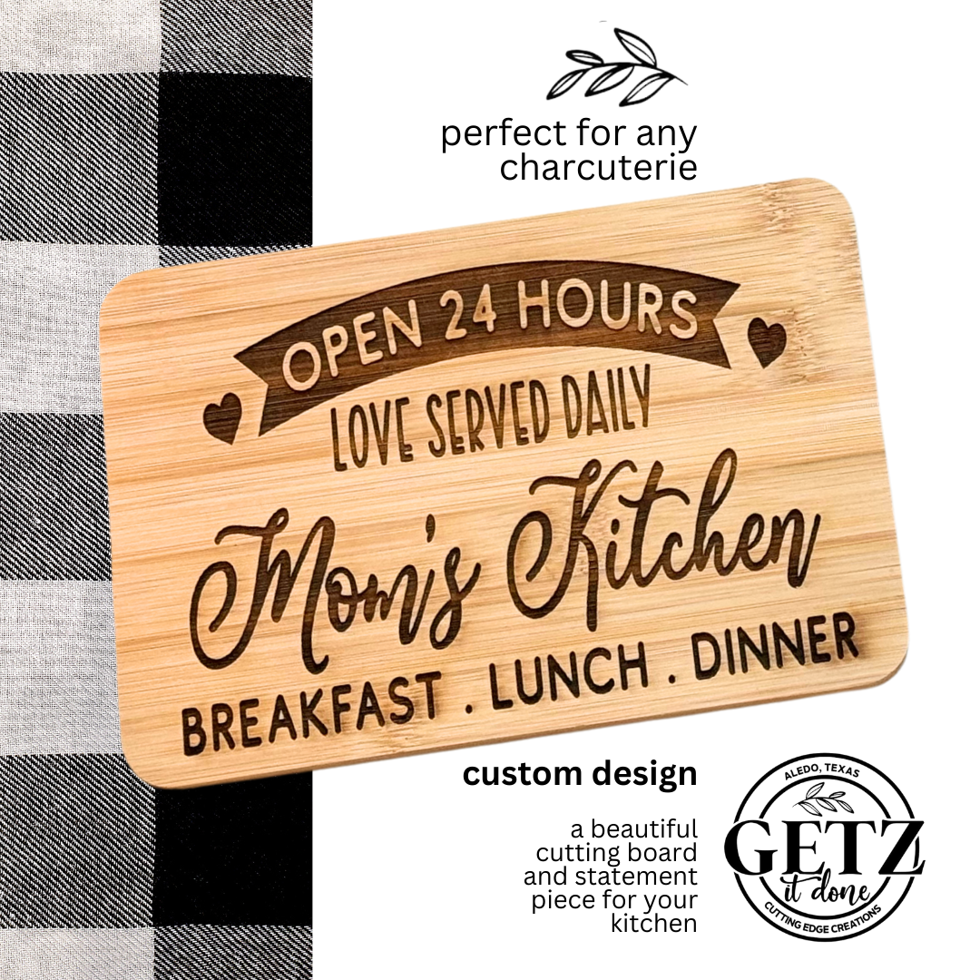 A wooden cutting board with the text Open 24 Hours, Love Served Daily, Moms Kitchen, Breakfast, Lunch, Dinner engraved on it. Its placed on a black and white checkered fabric. The board is described as perfect for charcuterie, and theres a Getz It Done logo on the bottom right.
