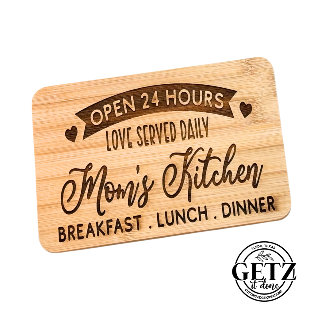 Rectangular wooden board with engraved text: Open 24 Hours, Love Served Daily, Moms Kitchen, Breakfast • Lunch • Dinner. Heart symbols decorate the text. A circular logo at the bottom right reads GETZ it done, Alvarado, Texas, Custom Edge Creations.