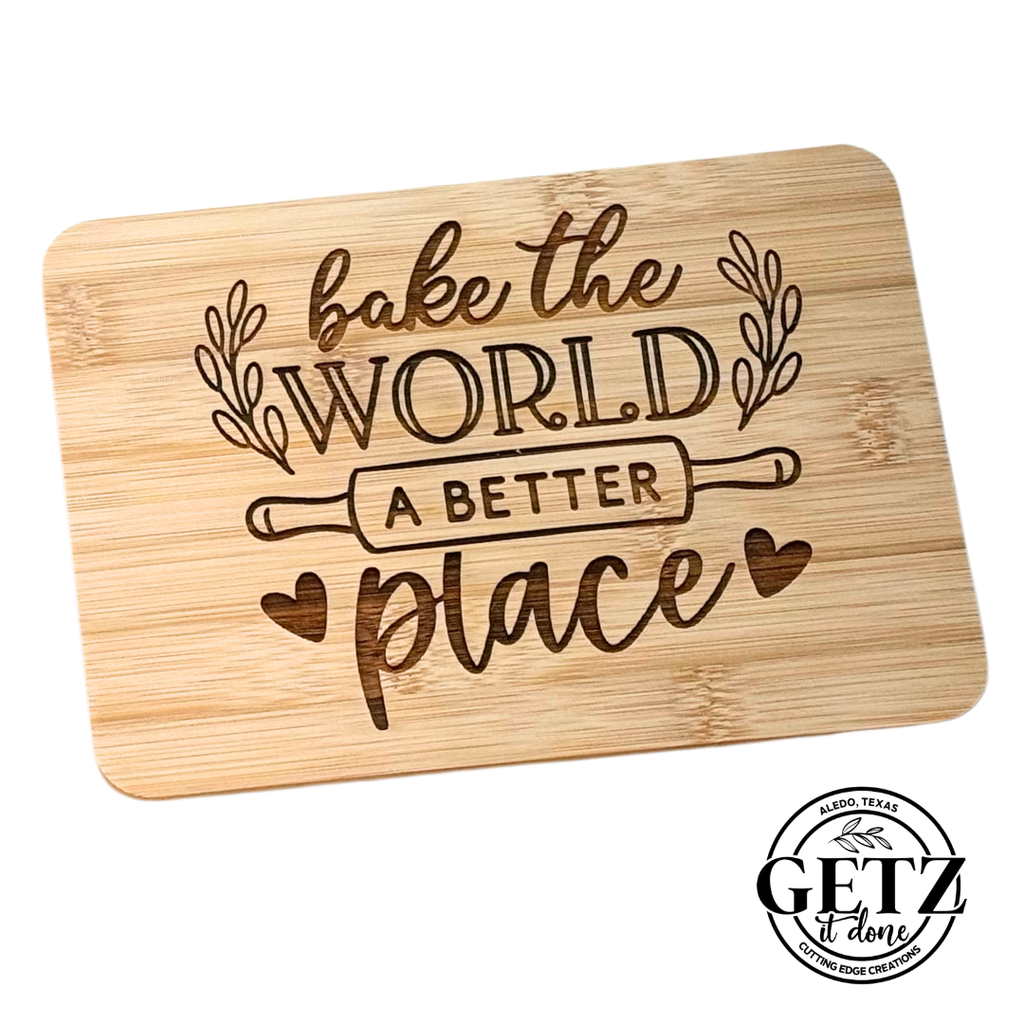 A wooden cutting board with engraved text reading Bake the World a Better Place surrounding an illustration of a rolling pin. Decorative leaves and hearts embellish the design. The bottom right corner features a round black and white logo with the text GETZ It done.