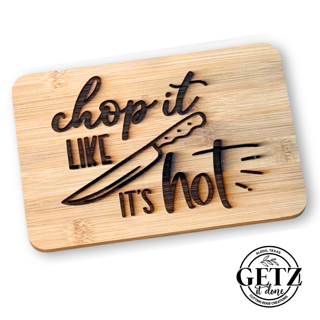 Cutting board with the phrase chop it like its hot engraved. Features a detailed illustration of a kitchen knife. Design is engraved on light wood. A round logo at the bottom right reads GETZ done - Cutting Edge Creations - Alamo, Texas.