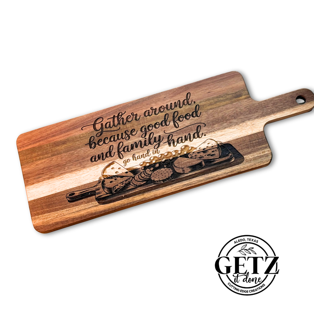 A rectangular wooden cutting board with a handle is engraved with the phrase Gather around, because good food and family go hand in hand. Below the text is an illustration of various cheeses and crackers. A circular logo in the corner reads GETZ it done, Alvord, Texas, Cutting Edge Creations.