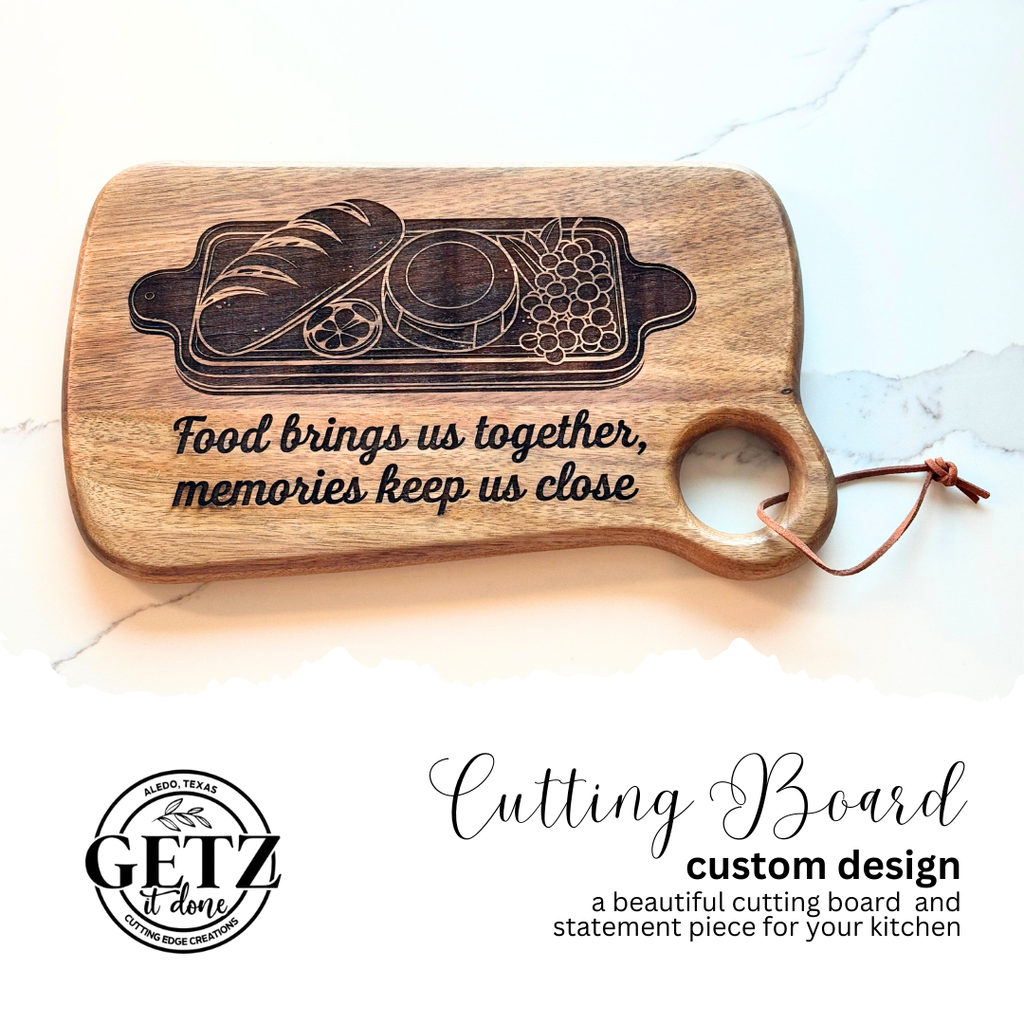 A wooden cutting board with an engraved design featuring bread, a jar, and grapes. Text reads, Food brings us together, memories keep us close. It has a round handle with a leather strap. Additional text below describes it as a custom design by Getz It Done.