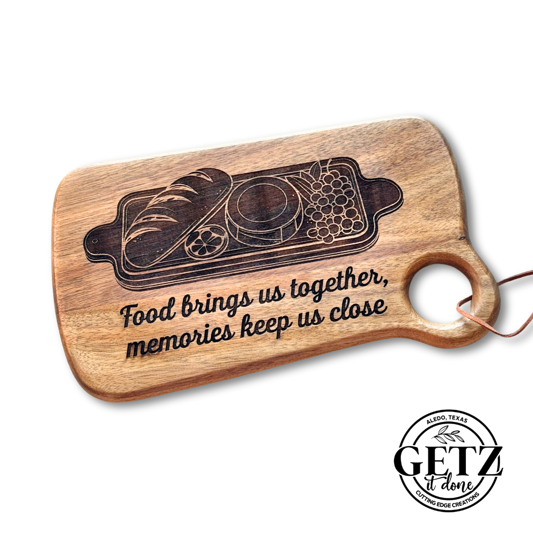 Wooden cutting board with a handle and leather loop. Engraved design includes bread, cheese, grapes, and a sliced tomato. Text reads, Food brings us together, memories keep us close. A GETZ done logo is in the bottom corner.