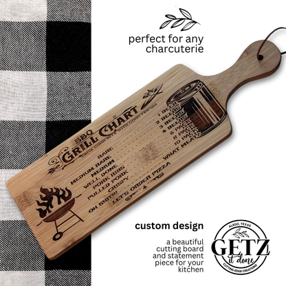 A wooden cutting board with a BBQ Grill Chart engraved, showing meat doneness levels from rare to well done. A grilling illustration and perfect for any charcuterie text are visible. The board has a handle, a leather strap, and rests on a black-and-white checkered cloth.
