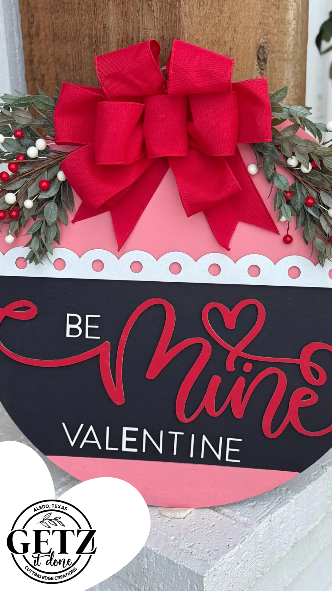 A round sign with a pink top and black bottom reads Be Mine Valentine in red and white text. Its adorned with a large red bow, green foliage, and small red and white berries. A logo in the corner says GETZ It Done.
