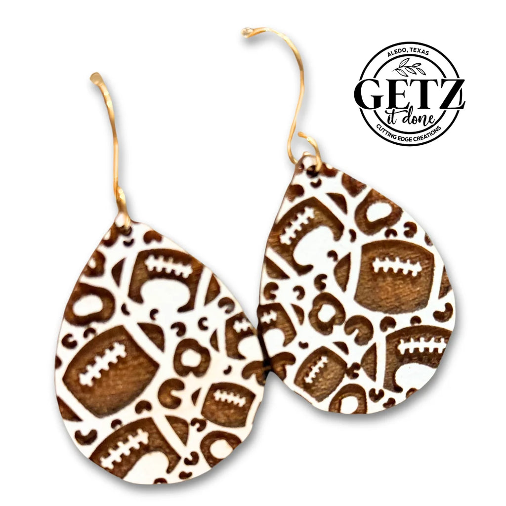 Teardrop-shaped earrings with a brown and white pattern. The design features football motifs and abstract shapes. Gold hooks are attached for wearing. A logo in the upper right corner reads “GETZ it done, Aledo, Texas, Cutting Edge Creations,” with a saw blade and leaves graphic.