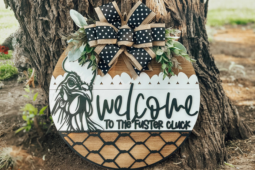 A round wooden sign with a chicken illustration, greenery, and a black-and-white polka dot bow reads “Welcome to the Fuster Cluck” in playful, bold lettering, leaning against a tree outdoors.