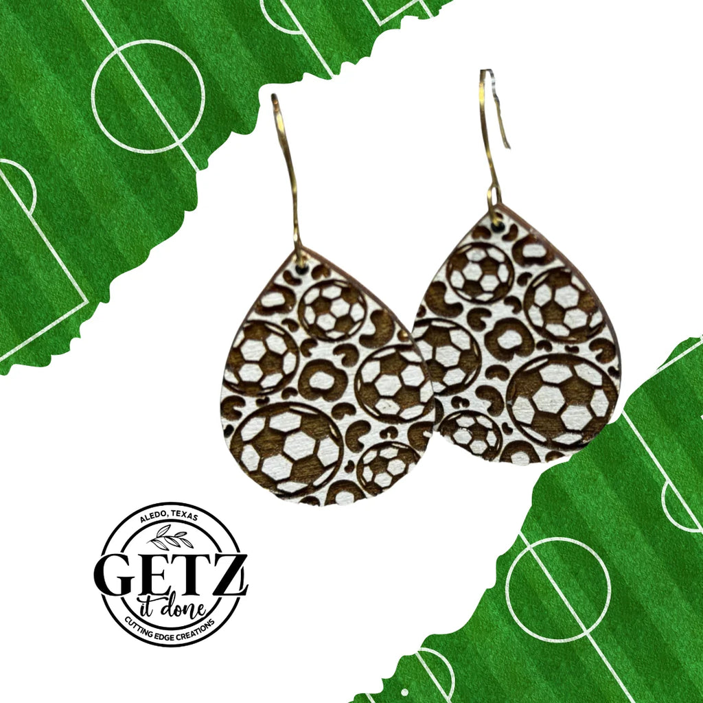 Teardrop-shaped earrings with a pattern of brown and white soccer balls. Positioned against a green background with white soccer field markings. The text GETZ it done and ALPRED, TEXAS CUTTING EDGE CREATIONS is seen near the bottom left.
