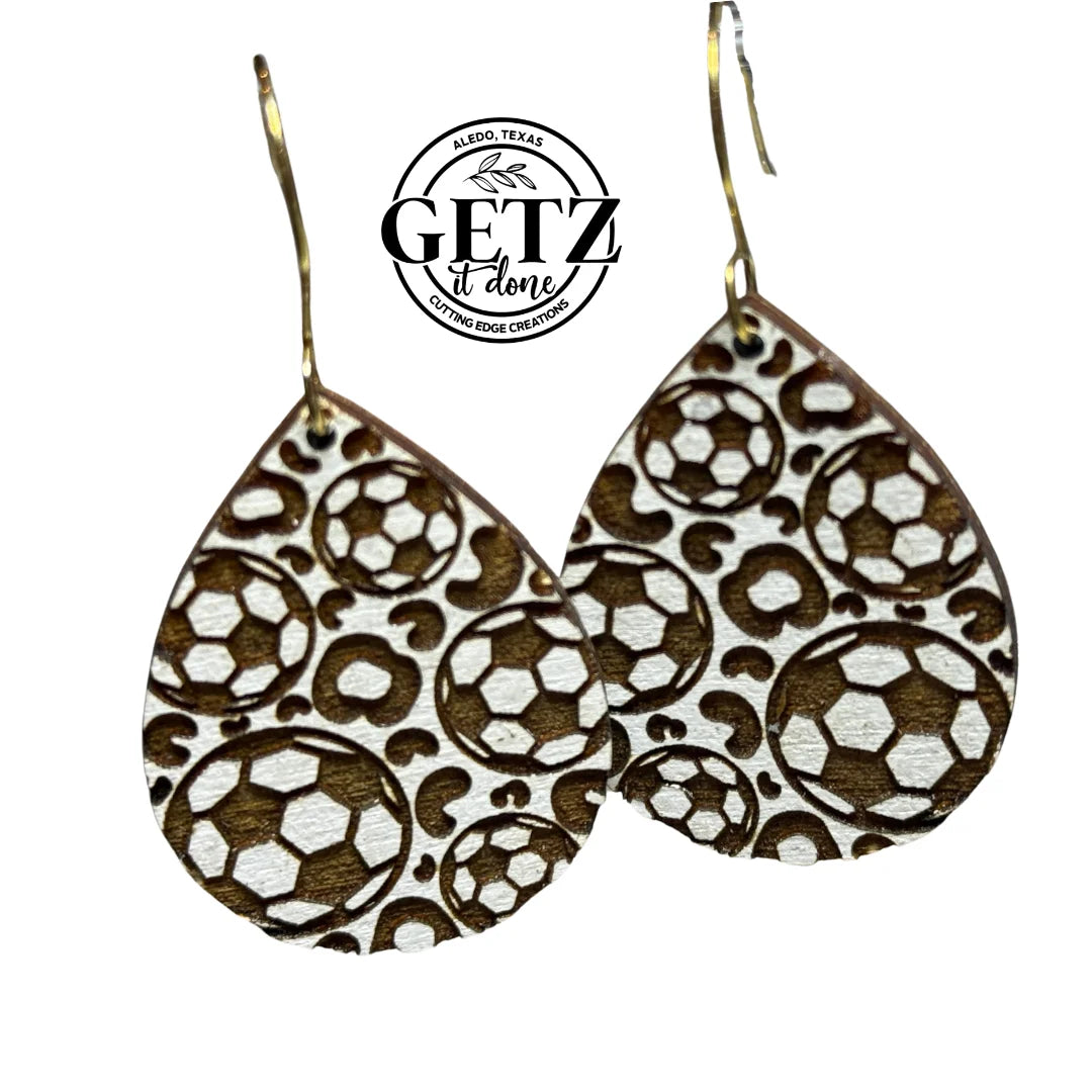 Teardrop-shaped earrings with a soccer ball pattern, featuring black and white hexagons. Above them is a circular logo with the text Aledo, Texas and Getz It Done Cutting Edge Creations. The earrings hang from thin metal hooks.