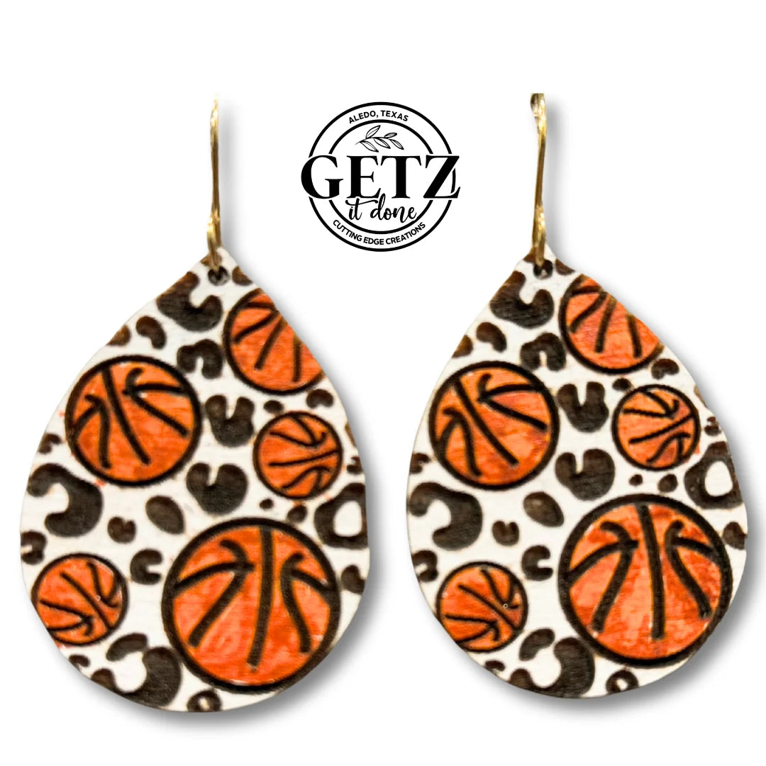 Teardrop-shaped earrings feature a leopard print background with orange and black basketball designs. A small circular logo at the top indicates GETZ it done, with text about crafting or creation. The hooks are metallic, contrasting with the vibrant patterns.