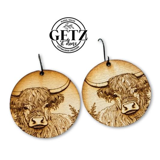 Round wooden earrings featuring engraved highland cow faces with distinctive long horns and shaggy fur. Above, a circular logo reads Getz It Done with Aledo, Texas and Cutting Edge Creations in smaller text, accompanied by a stylized feather.