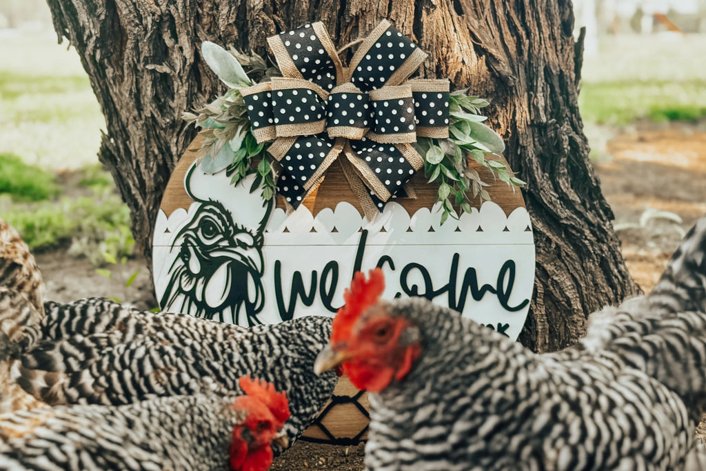 A round wooden sign with a chicken illustration, polka-dot bow, and greenery, reads welcome. Several black and white chickens with red combs stand in front of the sign, near a tree outdoors.