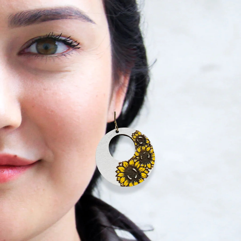 Close-up of a person wearing a circular earring with dried sunflowers embedded on one side. The earring has a central cutout. The persons skin is fair, with dark hair and visible eye, and they have a subtle smile. The background is blurred and light gray.