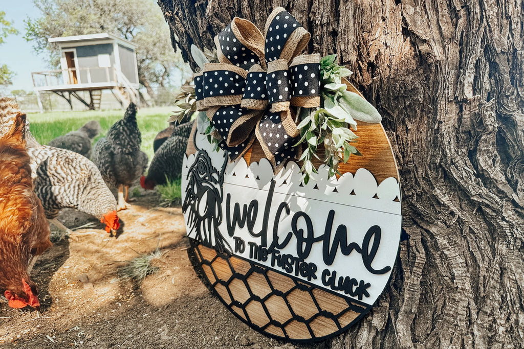 A decorative round sign with a bow and greenery reads Welcome to the Foster Cluck, hanging on a tree. Several chickens roam nearby, and a chicken coop is visible in the background.