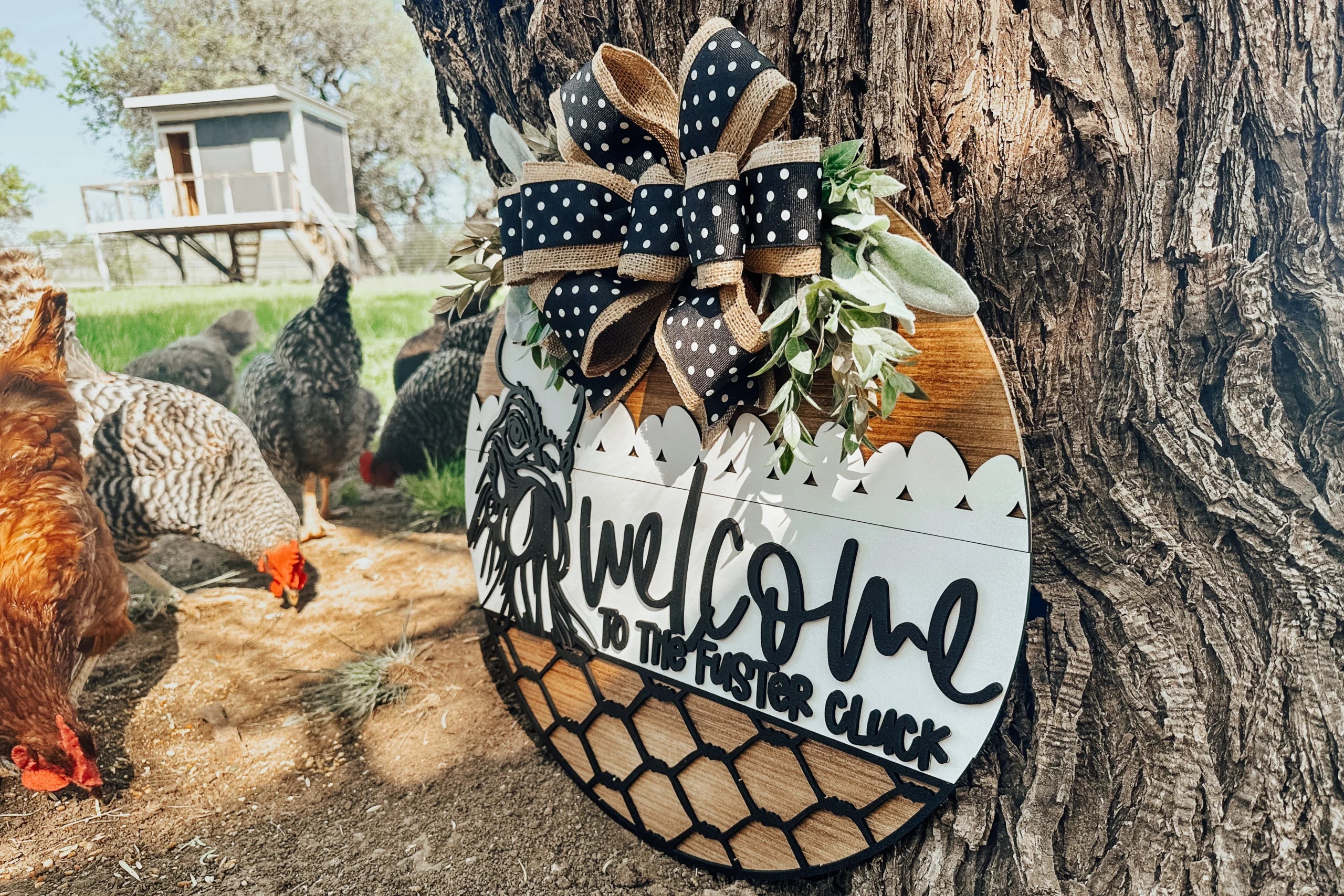 A decorative round sign with a bow and greenery reads Welcome to the Foster Cluck, hanging on a tree. Several chickens roam nearby, and a chicken coop is visible in the background.