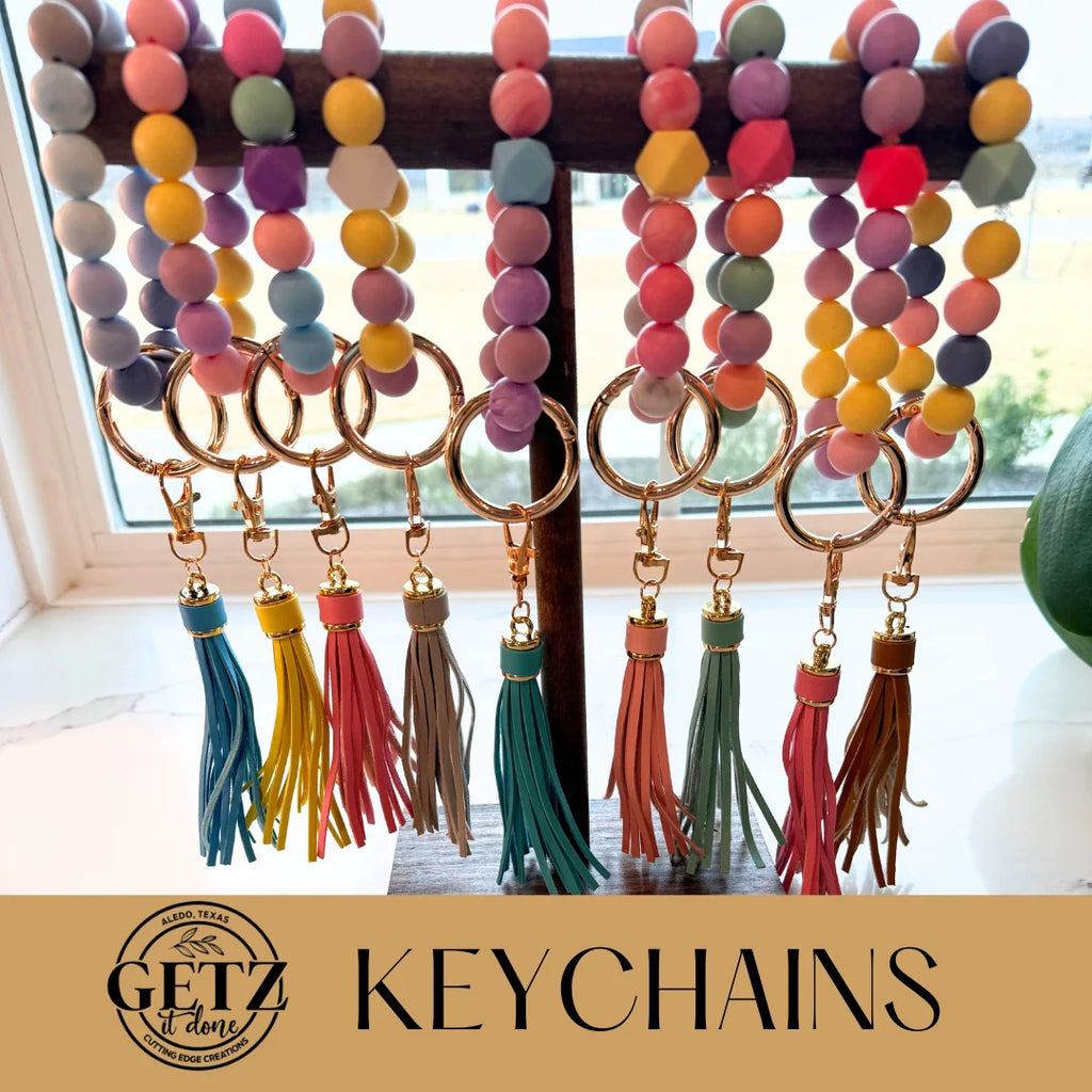 Colorful beaded keychains with tassels hanging on a wooden stand. The beads are round and hexagonal in shades of pink, blue, yellow, purple, and green. Gold rings and clasps hold assorted tassels in pink, gray, teal, yellow, and red. The text Getz it done and Keychains is displayed below.