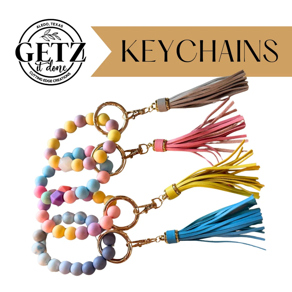 Four colorful keychains with round beads and tassels are displayed diagonally. The beads are in pastel shades, and each keychain features a gold ring. Tassels in tan, pink, yellow, and blue accompany the beads. Text reads Getz it Done Keychains with a logo saying Aledo, Texas Cutting Edge Creations.