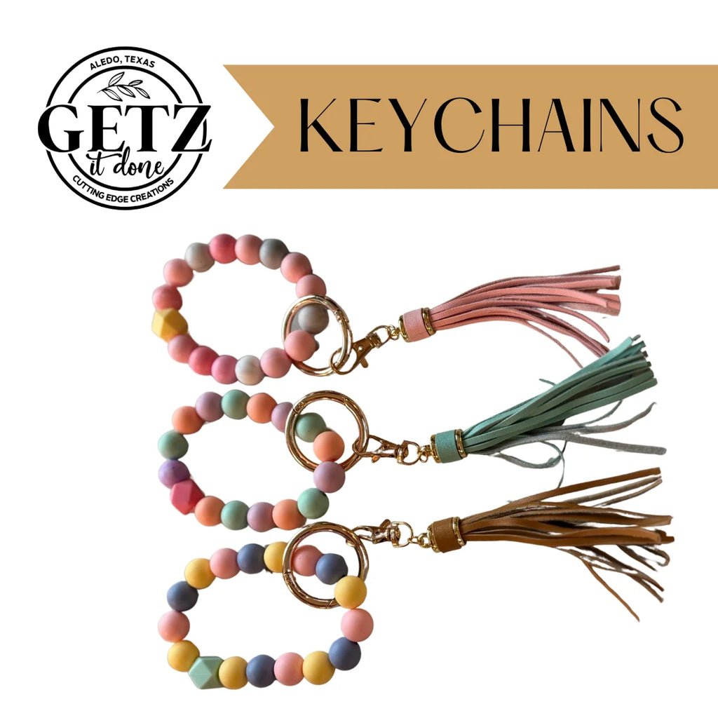 Three colorful keychains with round and geometric beads are displayed. Each has a tassel—pink, green, and brown—and a gold clasp. A logo with GETZ It Done in black text and KEYCHAINS on a brown banner is at the top.