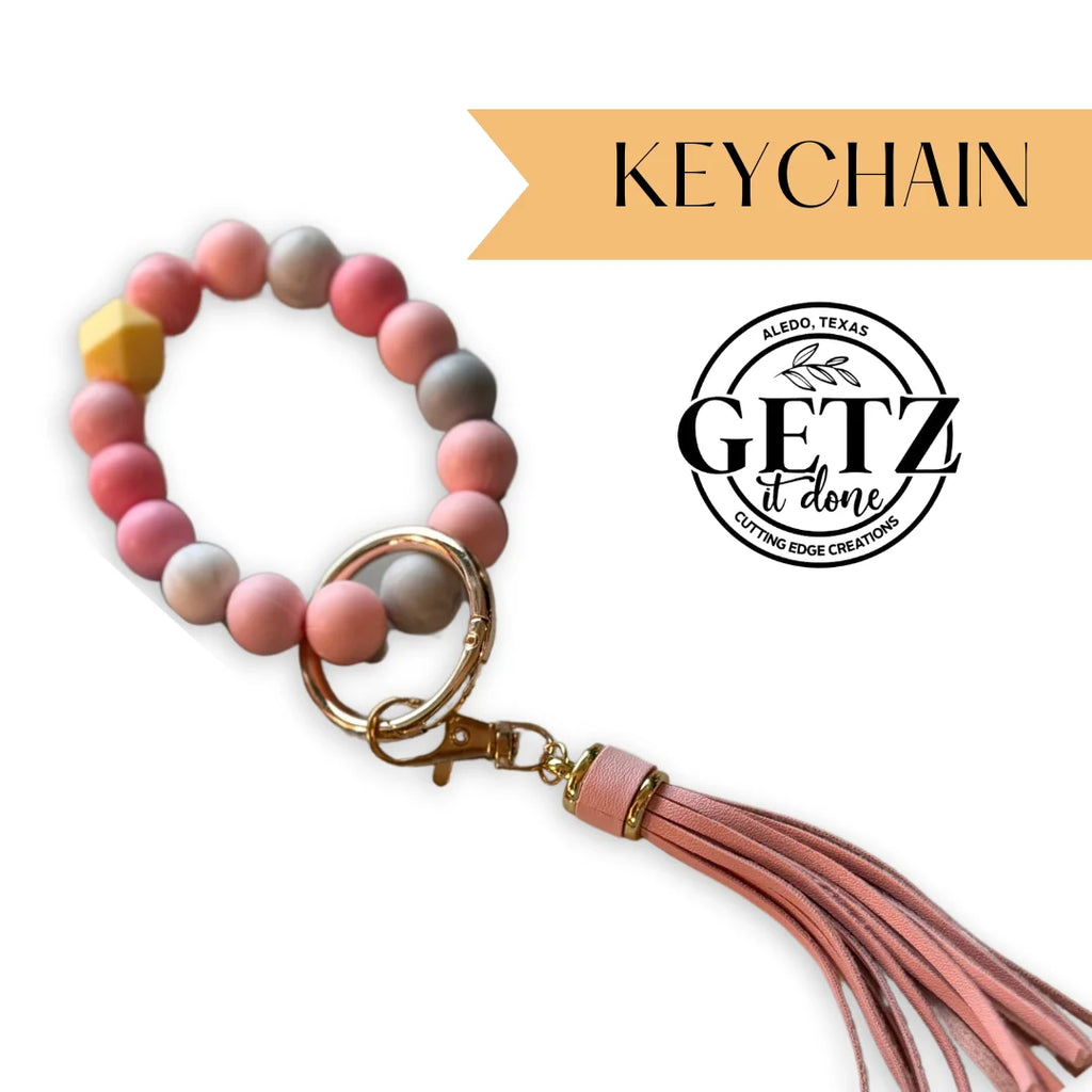 Keychain with pink, gray, and yellow beads on a loop, featuring a gold clasp and a pink leather-like tassel. Background includes a logo with Getz It Done and the text Aledo, Texas Cutting Edge Creations, along with a yellow banner labeled KEYCHAIN.