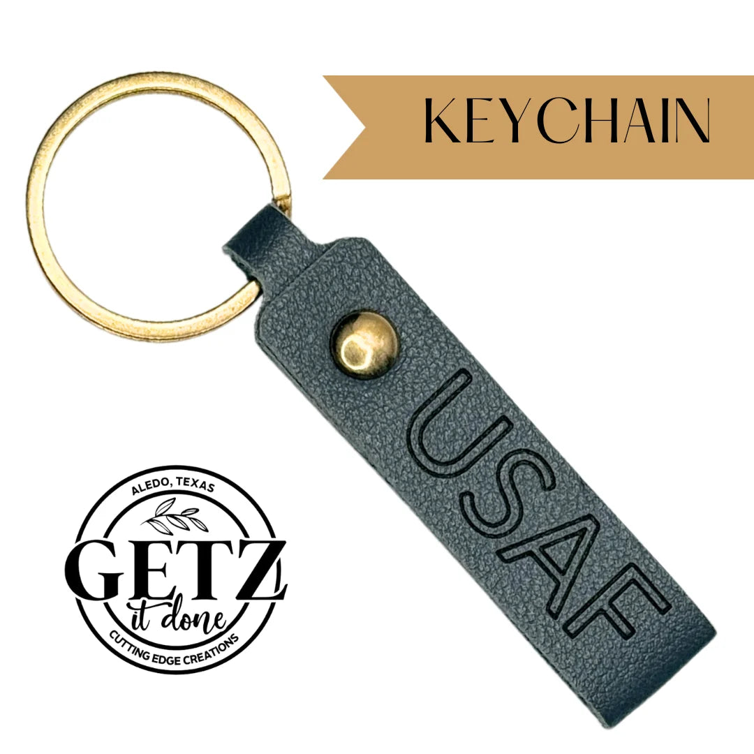 Gold keychain with a dark gray leather strap embossed with USAF. The strap has a gold snap button and is attached to a circular ring. The design includes a logo reading Aledo, Texas, Getz It Done, Cutting Edge Creations in black text on a white background.