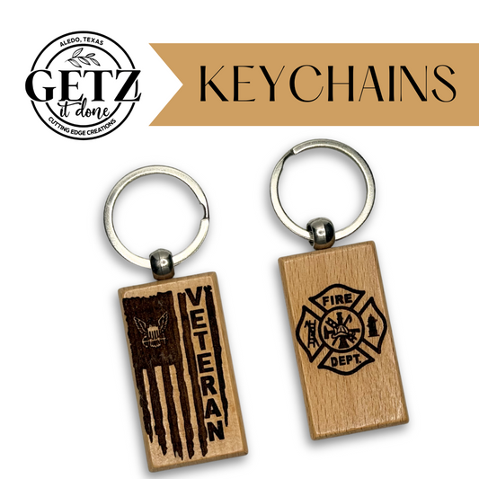 Two wooden keychains with silver rings. The left keychain has an engraved American flag with a skull and the word VETERAN. The right keychain features a fire department emblem with crossed axes and a helmet. The background includes a logo and the word KEYCHAINS in a beige banner.