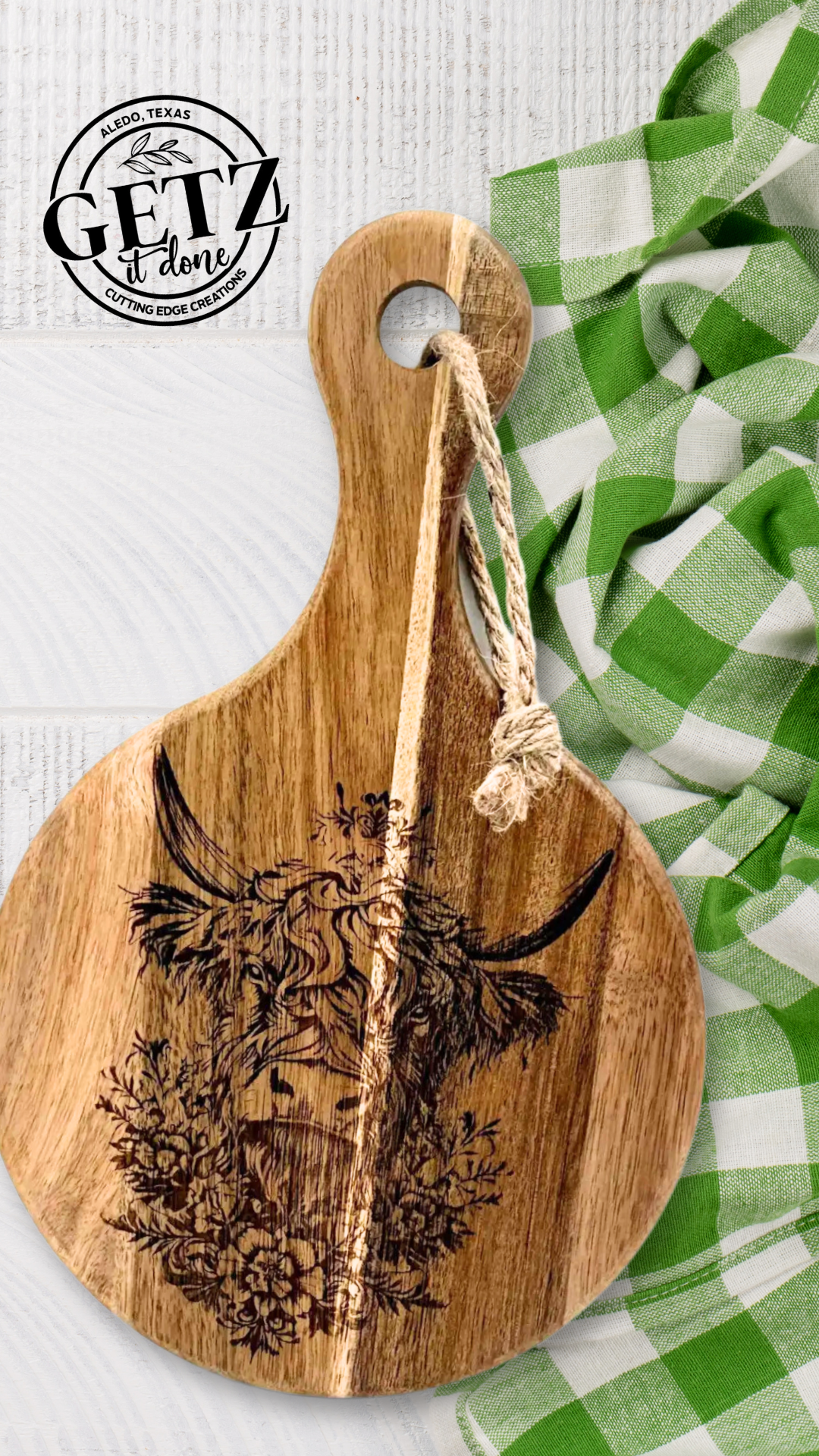Round wooden cutting board with an engraved image of a highland cow surrounded by flowers. A rope loop is attached to the handle. It rests on a white wood surface beside a crumpled green and white checkered cloth. A circular logo at the top says Getz it done with Aledo, Texas.