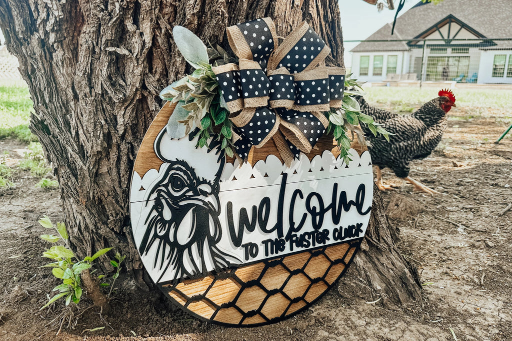 A round wooden sign leaning against a tree reads Welcome to the Flock with a chicken illustration, decorative ribbon, faux greenery, and chicken wire details. A black-and-white chicken stands nearby on the grass.