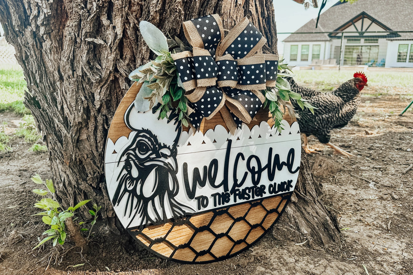 A round wooden sign leaning against a tree reads Welcome to the Flock with a chicken illustration, decorative ribbon, faux greenery, and chicken wire details. A black-and-white chicken stands nearby on the grass.