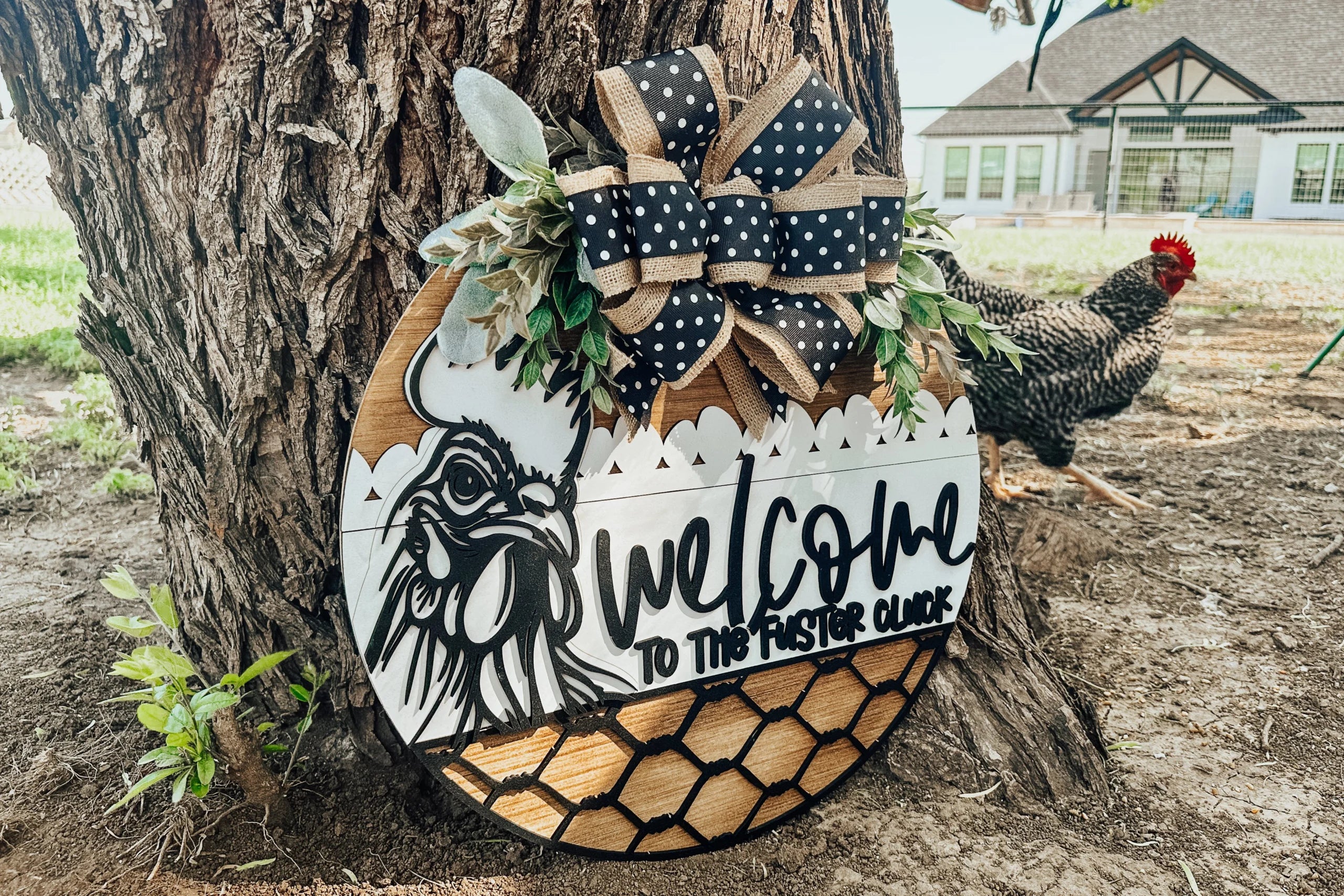 A round wooden sign leaning against a tree reads Welcome to the Flock with a chicken illustration, decorative ribbon, faux greenery, and chicken wire details. A black-and-white chicken stands nearby on the grass.