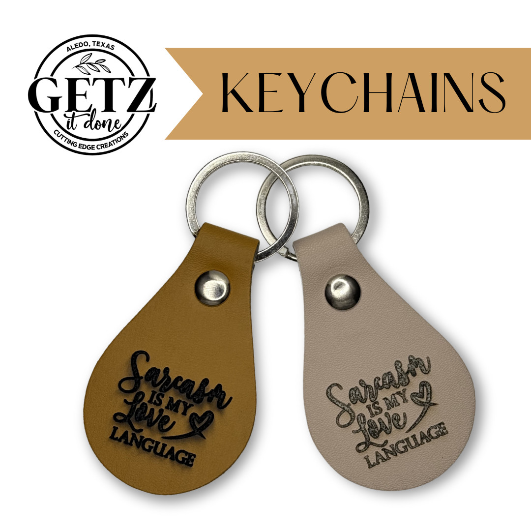 Two teardrop-shaped leather keychains with metal rings, one tan and one gray. Both have Sarcasm is my love language engraved in black. The top left features the GETZ it done logo from Aledo, Texas. The text KEYCHAINS is on a beige banner above.