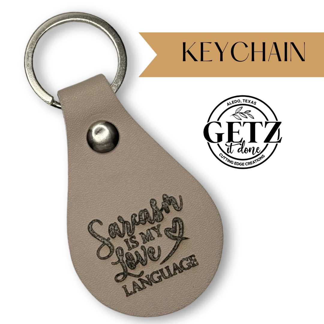 Leather teardrop keychain with engraved text Sarcasm is my love language and an arrow with a heart. Features a metal snap and ring. Background includes a logo with GETZ it done and Aledo, Texas Cutting Edge Creations. Tan label in the top right reads KEYCHAIN.