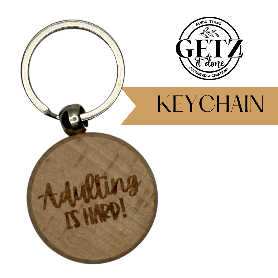 A wooden keychain with the engraved text Adulting is hard! in a casual font. Attached to a silver metal key ring. The top right shows the logo and text GETZ it done and KEYCHAIN on a brown banner. Located in Aledo, Texas, focus on custom creations.
