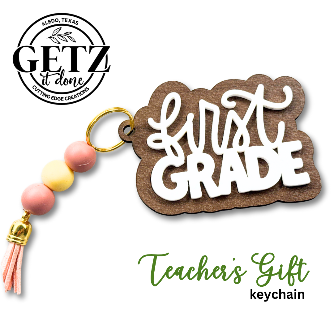 Keychain with a wooden First Grade plaque, white script text raised on top. Attached are three round beads in pink, yellow, and pink, and a pink tassel. GETZ It Done logo and text Teachers Gift keychain appear on the white background.