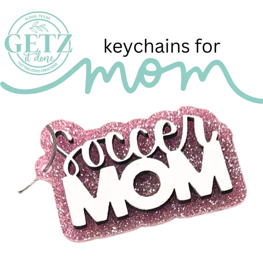 A sparkly pink keychain with the words Soccer MOM in bold white and black letters. Above it, the text keychains for mom is written in black and teal. The top left corner features the circular logo GETZ it done with Aledo, Texas and Cutting Edge Creations written within it.
