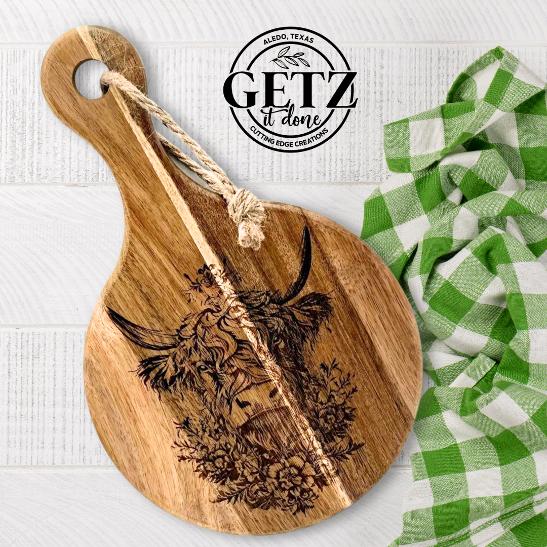 A wooden cutting board with an engraved cow and flowers leans against a white surface. A green and white checkered cloth is partially visible on the right. A rope is looped through the board’s handle. The logo, GETZ it done, Aledo, Texas, is displayed above.

.