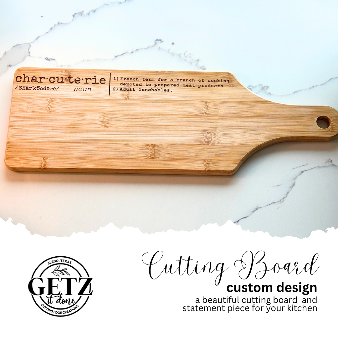A bamboo cutting board displays the definition of charcuterie printed on it: 1) French term for a branch of cooking, 2) devoted to prepared meat products, 3) adult lunchables. The board is set on a white marble surface. Below, black text reads Cutting Board custom design next to a logo.