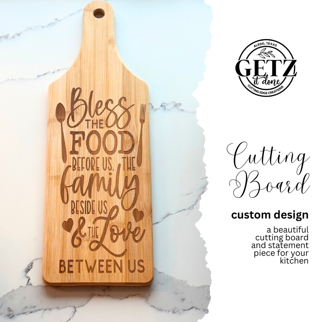 A wooden cutting board is engraved with the text Bless the food before us, the family beside us & the love between us. It features a hole for hanging. Next to it, the text describes it as a custom design and a statement piece for the kitchen. A circular logo is at the top right.