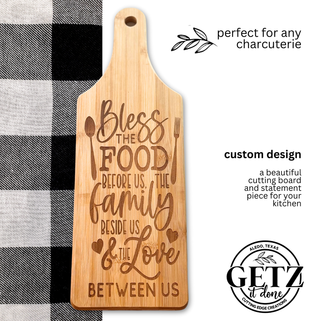 A wooden cutting board engraved with the phrase: Bless the food before us, the family beside us, & the love between us. Its styled with heart and utensil designs. The board is placed on a black and white checkered cloth, with text promoting it as perfect for any charcuterie.