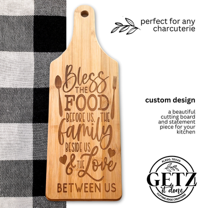 A wooden cutting board engraved with the phrase: Bless the food before us, the family beside us, & the love between us. Its styled with heart and utensil designs. The board is placed on a black and white checkered cloth, with text promoting it as perfect for any charcuterie.