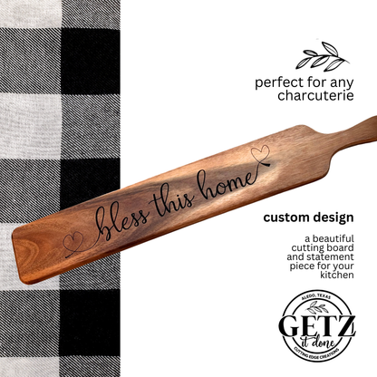 A wooden charcuterie board with bless this home written in cursive, featuring small heart motifs. Its positioned on a black and white checkered fabric. Text reads, perfect for any charcuterie, custom design, with GETZ done logo at the bottom.