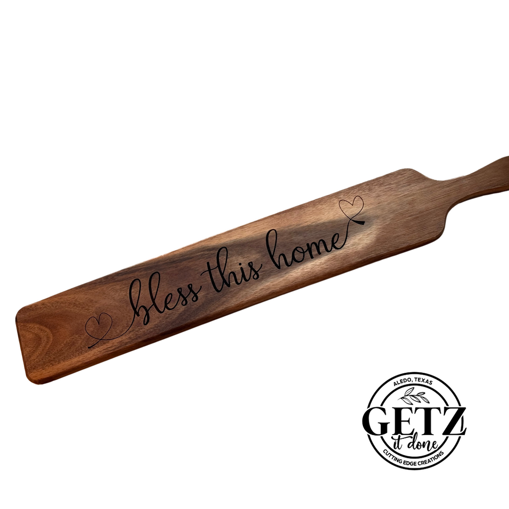 Wooden paddle with bless this home and heart designs engraved. The paddle has a smooth finish and a handle on one side. The logo GETZ it done, Aledo, Texas, Custom Engraving is in black at the bottom right corner.