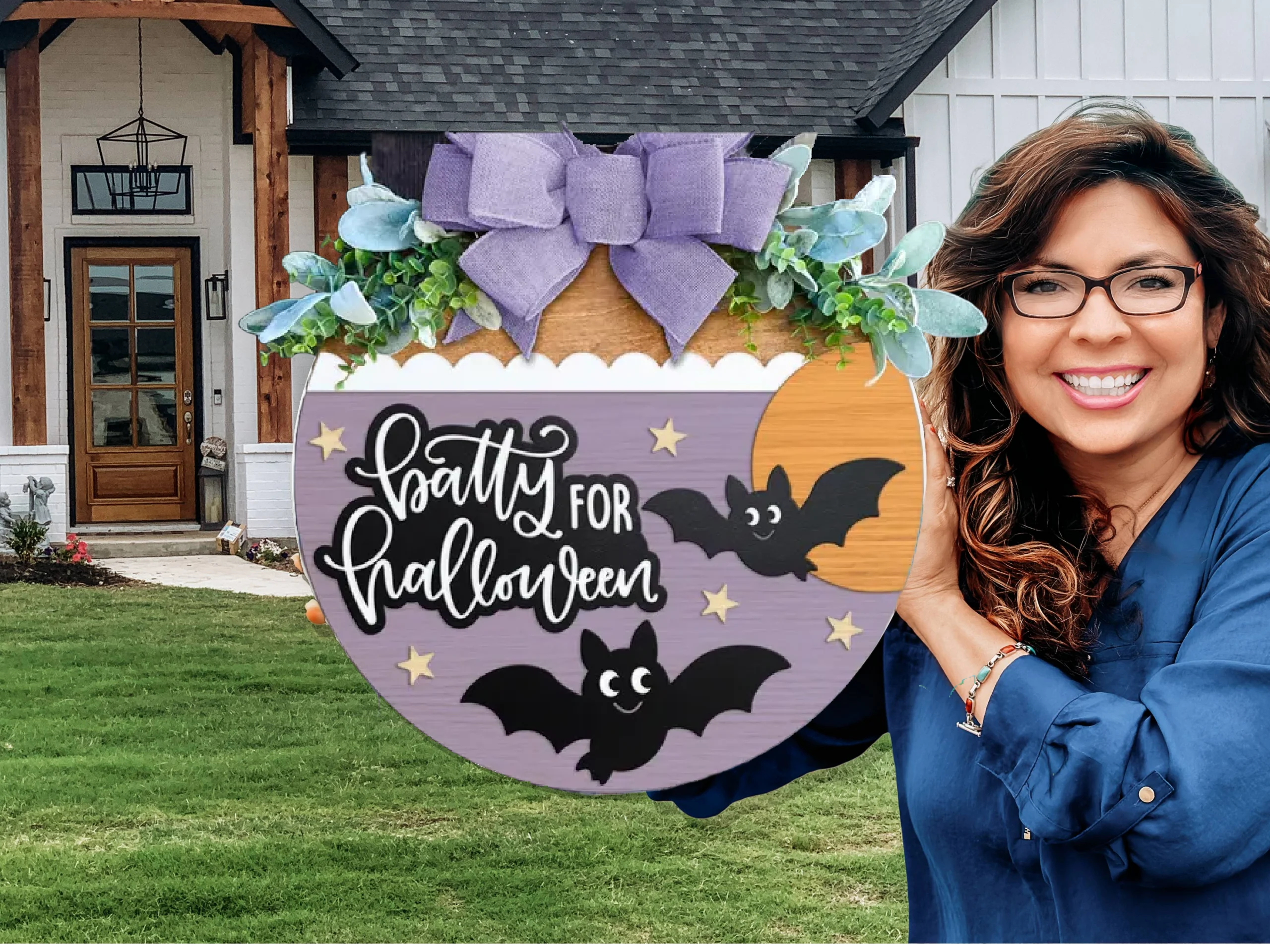 A smiling woman with wavy hair holds a round Halloween-themed sign decorated with bats, greenery, and a purple bow, reading Batty for Halloween, standing in front of a modern house with a lawn.