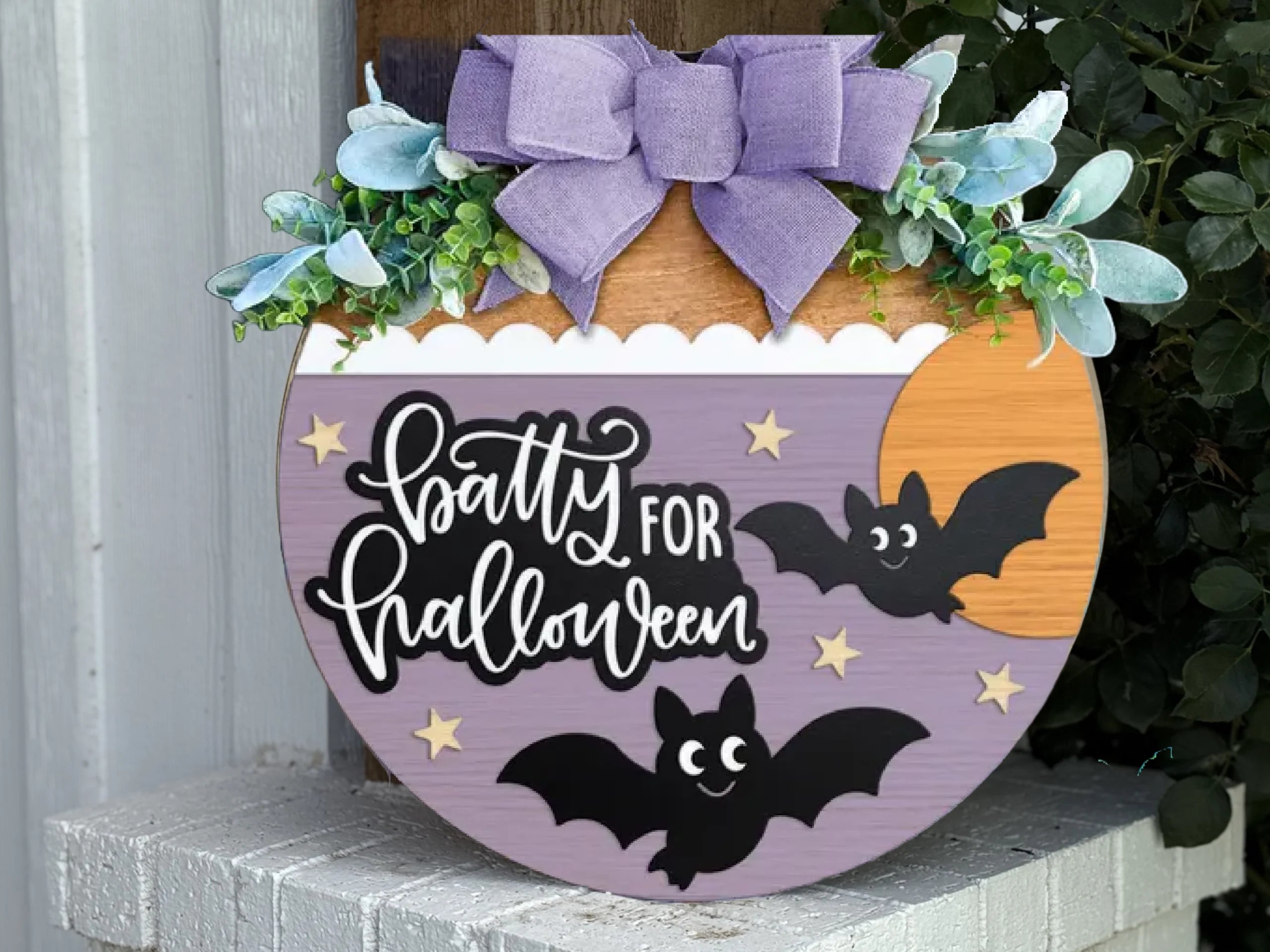 A round wooden Halloween sign decorated with a purple bow, greenery, and two cartoon bats. The sign reads Batty for Halloween and has stars and a yellow moon in the background.
