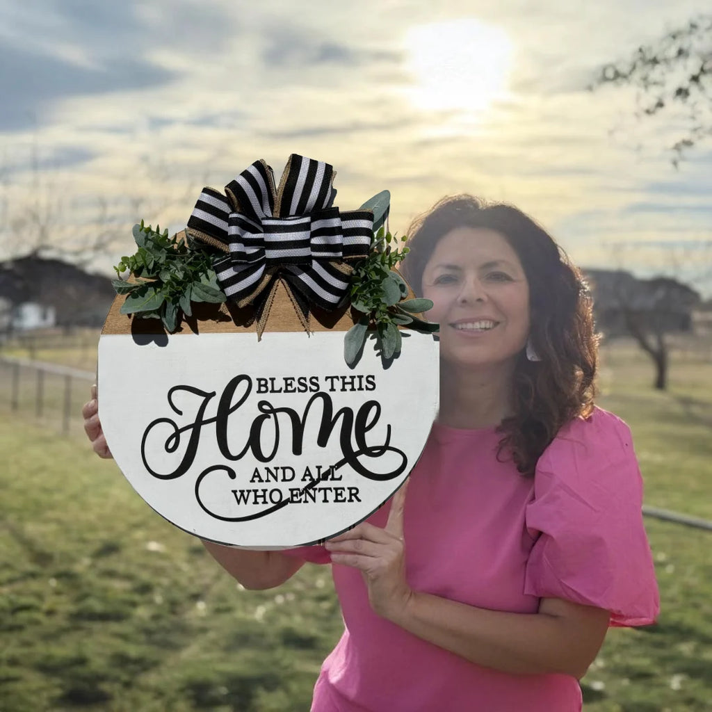 A woman in a pink blouse stands outdoors, smiling and holding a decorative circular sign. The sign features a black and white striped bow with green foliage at the top, and the message Bless this home and all who enter in elegant black script. The sky is partly cloudy.