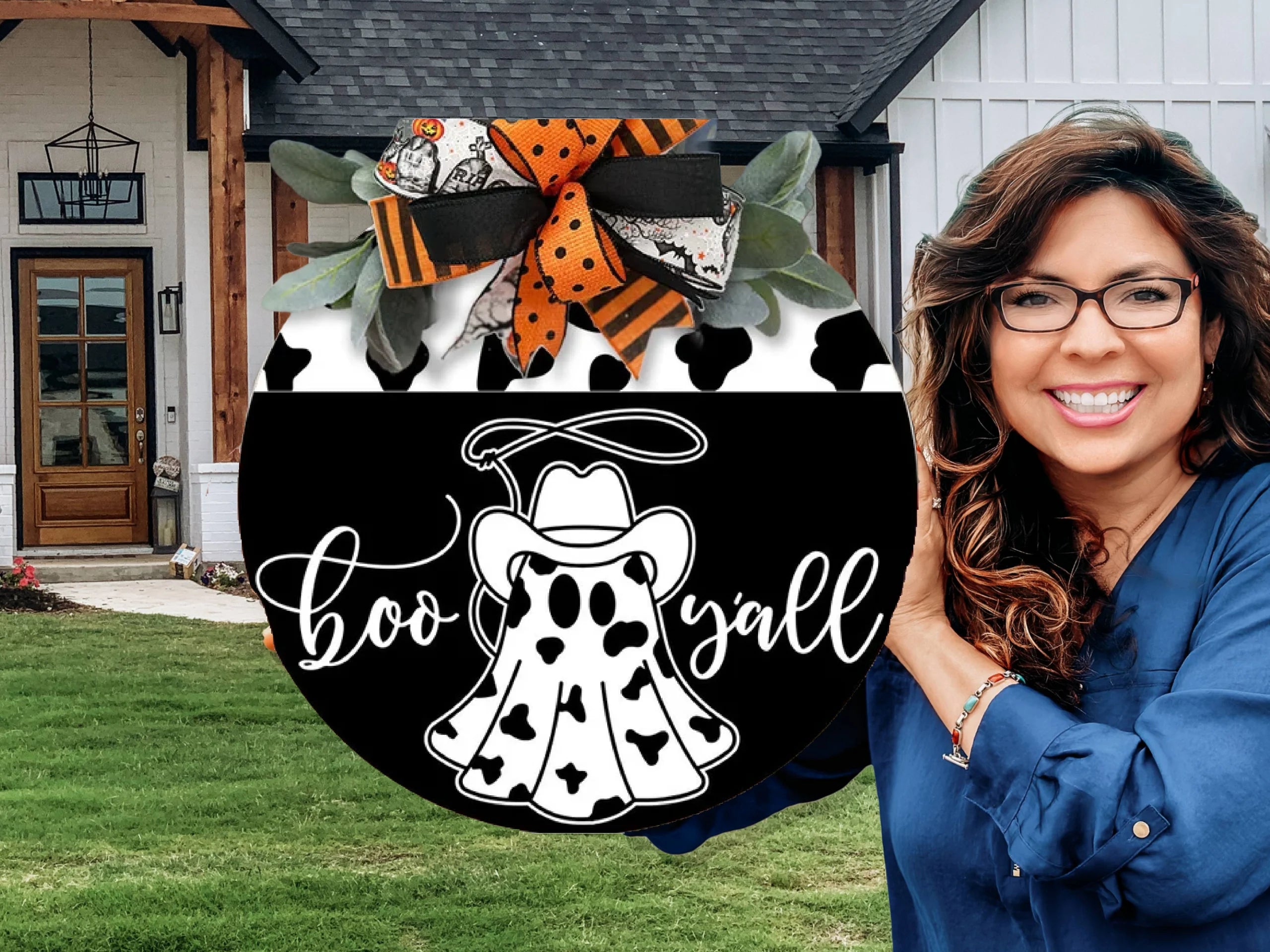 A smiling woman with wavy brown hair and glasses holds a round Halloween sign that says boo yall, featuring a ghost in a cowboy hat. The sign is decorated with orange and black ribbons. A house is in the background.