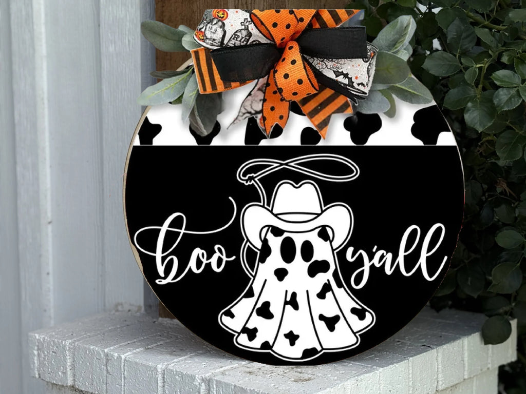 A round wooden sign features a ghost in a cowboy hat and cow-print cloak, with the words boo yall in white script. The sign is decorated with orange, black, and white bows and greenery at the top.