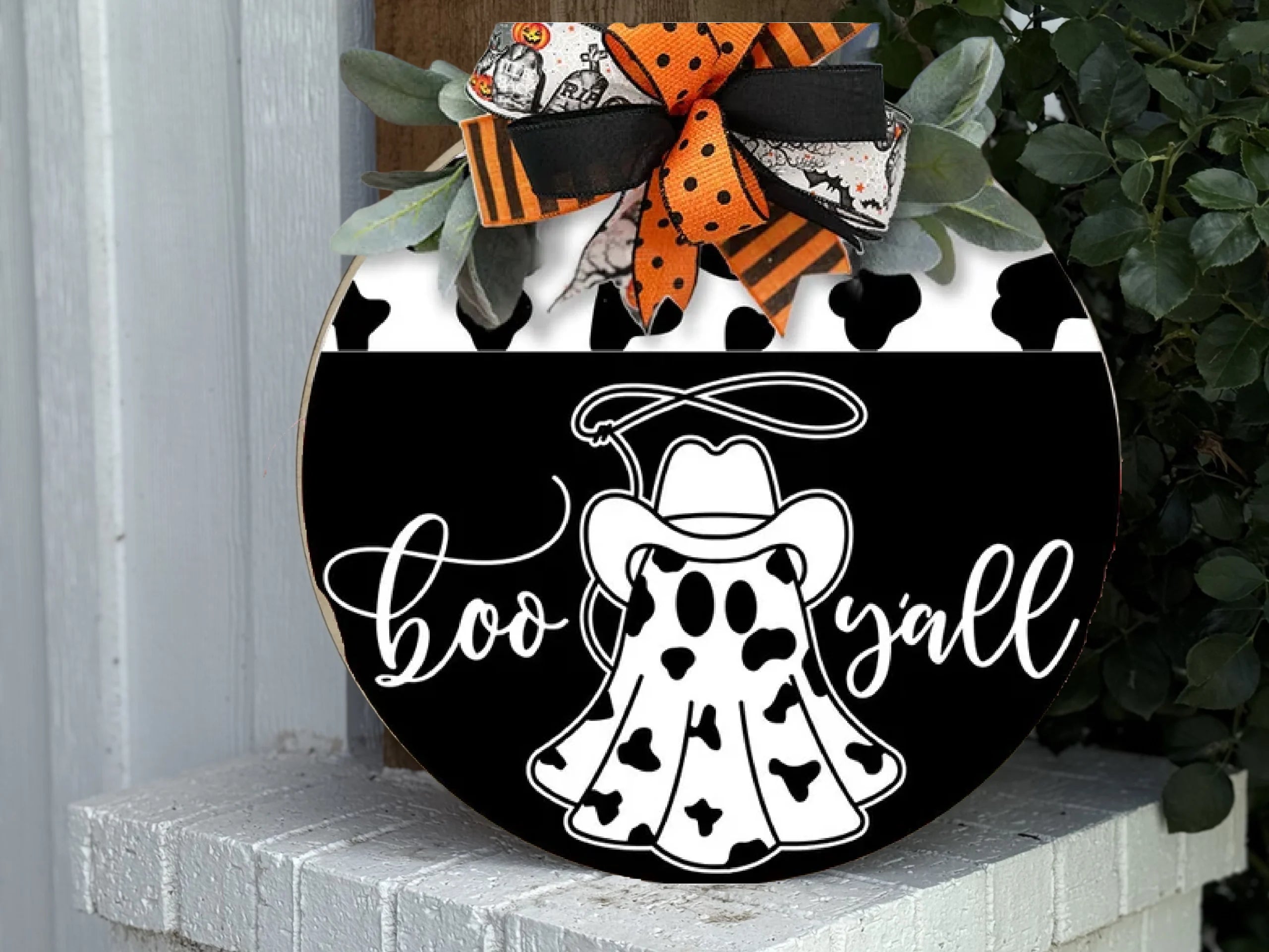A round wooden sign features a ghost in a cowboy hat and cow-print cloak, with the words boo yall in white script. The sign is decorated with orange, black, and white bows and greenery at the top.