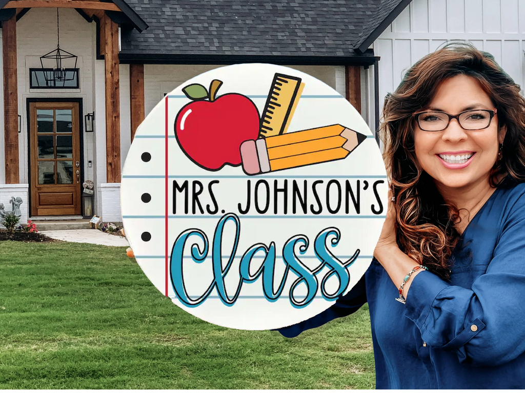 A smiling woman with long brown hair and glasses stands outside a house, holding a round sign with an apple, pencils, and ruler that reads Mrs. Johnsons Class.